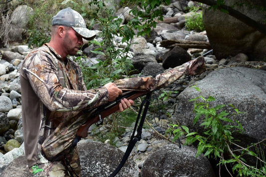 A man in camouflage holds a rifle in the woods, equipped with a waterproof gun cover for protection against the elements.